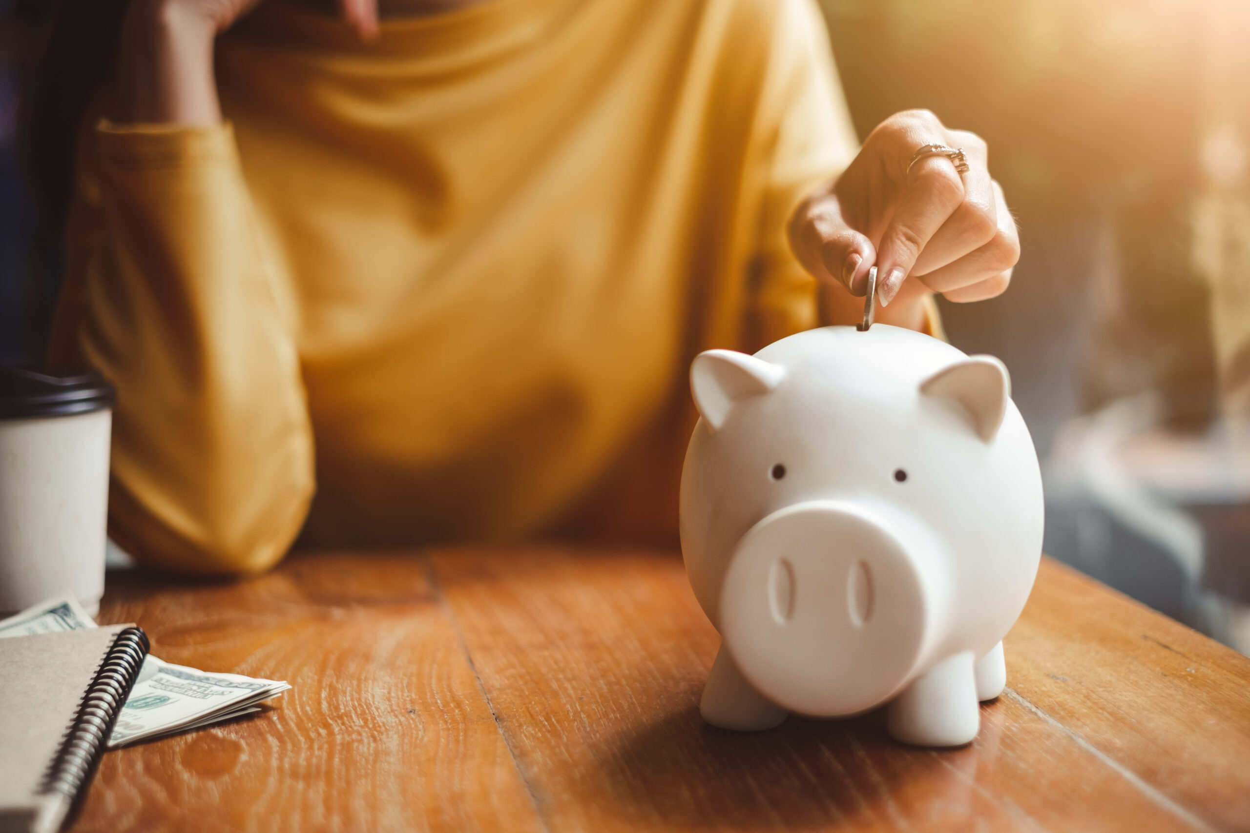 Person in a yellow sweater saving money by placing a coin into a white piggy bank on a wooden table, symbolizing student discounts in Bolton.